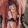 baby, child, stroller, pink, headband, bow, infant, seated, safety_harness, dress, floral_pattern, cute, indoors, carriage, person, portrait, wide_eyes, curious, soft_lighting, baby_clothing