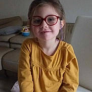 Liana participe au concours pour gagner de l'argent avec cette photo : child, girl, glasses, yellow_shirt, long_sleeves, smile, sitting, chair, living_room, couch, pillow, toy, floor, hair, indoor, person, casual_clothing, face, eyewear, relaxed