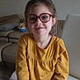 child, girl, glasses, yellow_shirt, long_sleeves, smile, sitting, chair, living_room, couch, pillow, toy, floor, hair, indoor, person, casual_clothing, face, eyewear, relaxed