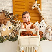 Ayaz a rejoint le concours — aidez-le/la à gagner de superbes lots ! toddler, toy_jeep, bow_tie, suspenders, plush_giraffe, stuffed_toy, miniature_house, decor, child, smile, indoor, playful, cute, white_floor, brick_wall, orange, brown, greenery, photography, portrait