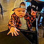 Charlie is registered to the contest to win money with this photo: child, baby, toddler, cap, backwards_cap, plaid_shirt, flannel_shirt, orange_black, highchair, table, hand, fingers, indoor, restaurant, seat, tray, expression, face, stroller, person