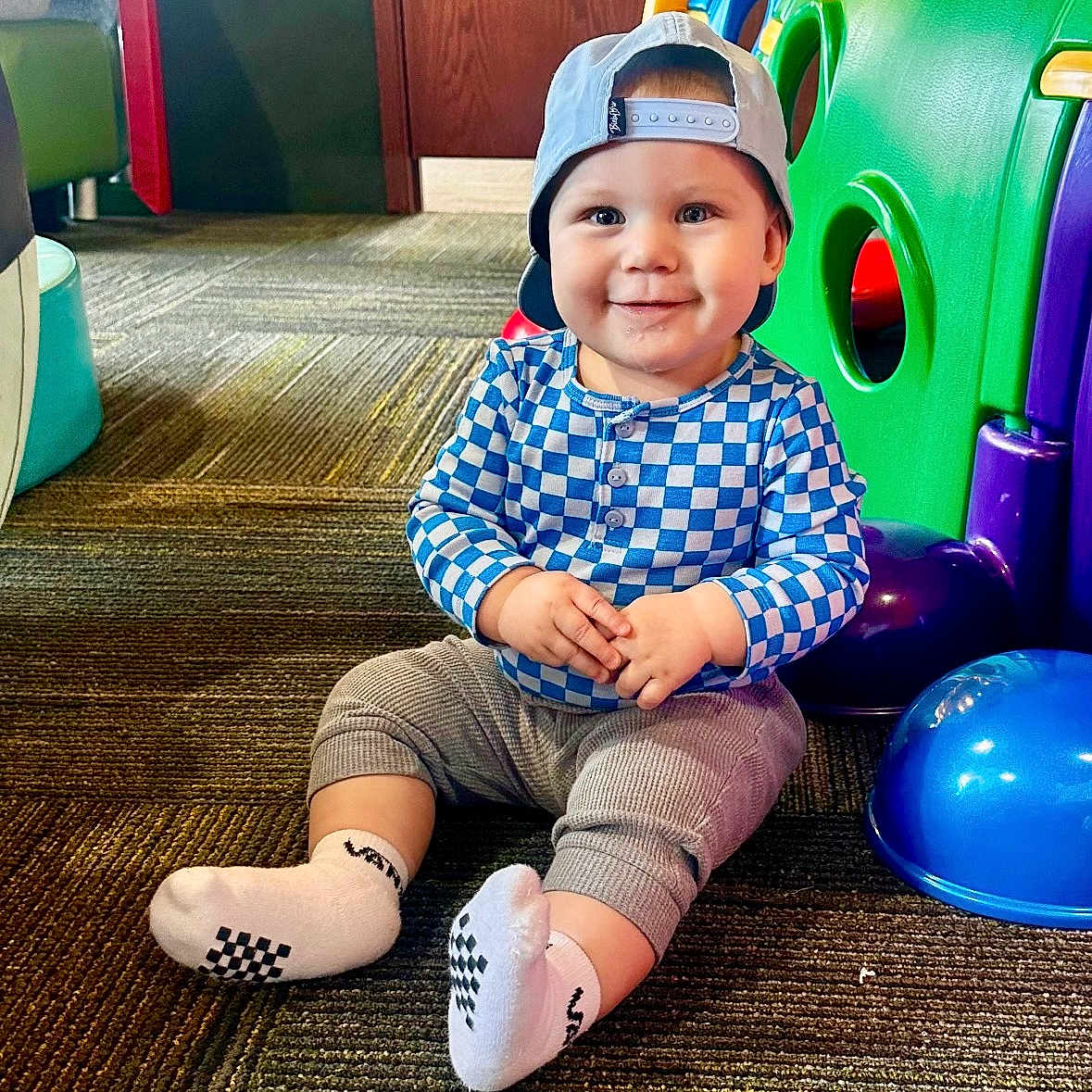 Charlie is registered to the contest to win money with this photo: baby, backwards_hat, cap, carpet, checkered_shirt, child, chubby_cheeks, colorful, cute, floor, happy, indoor, plastic_tunnel, play_area, portrait, sitting, smile, socks, toddler, toy