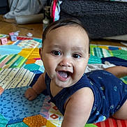 Caleb is registered to the contest to win money with this photo: baby, smiling, play_mat, indoor, child, colorful, floor, happy, infant, crawling, cute, pattern, toy, person, home, young_child, bright, cheerful, blue_clothing, closeup