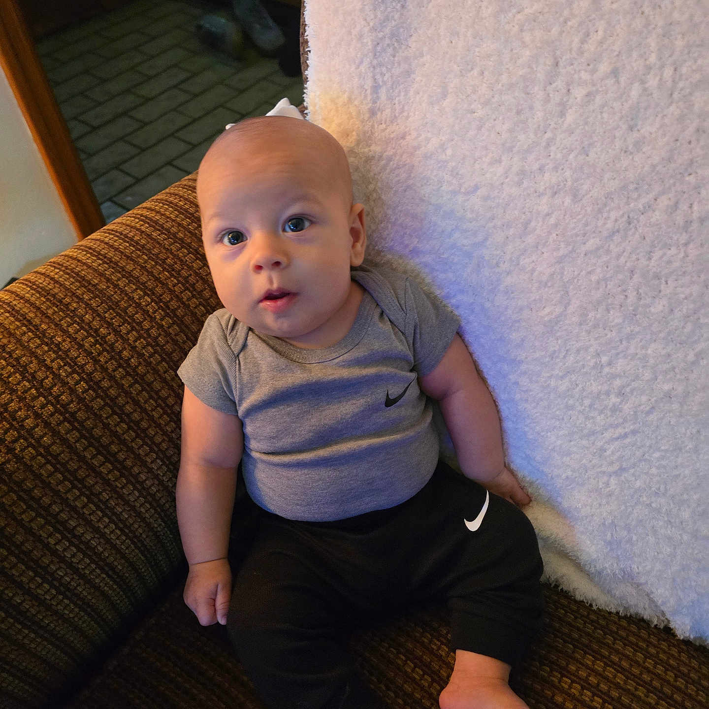 Kaysin joined the competition — help win amazing prizes! baby, barefoot, black_pants, carpeted_floor, child, clothing, couch, curious_expression, footwear_absent, grey_shirt, home_interior, indoors, infant, looking_up, nike_logo, patterned_fabric, portrait, sitting, soft_blanket, wooden_furniture