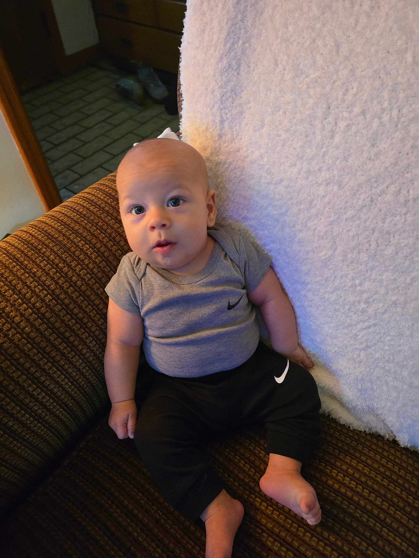 Kaysin joined the competition — help win amazing prizes! baby, infant, child, couch, clothing, grey_shirt, black_pants, nike_logo, sitting, indoors, footwear_absent, patterned_fabric, curious_expression, looking_up, barefoot, soft_blanket, home_interior, wooden_furniture, carpeted_floor, portrait