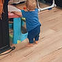toddler, child, blue_clothing, barefoot, wooden_floor, plastic_table, indoor, toy_scooter, high_chair, standing, back_view, home, flooring, young_child, furniture, person, baby, support, play_area, casual