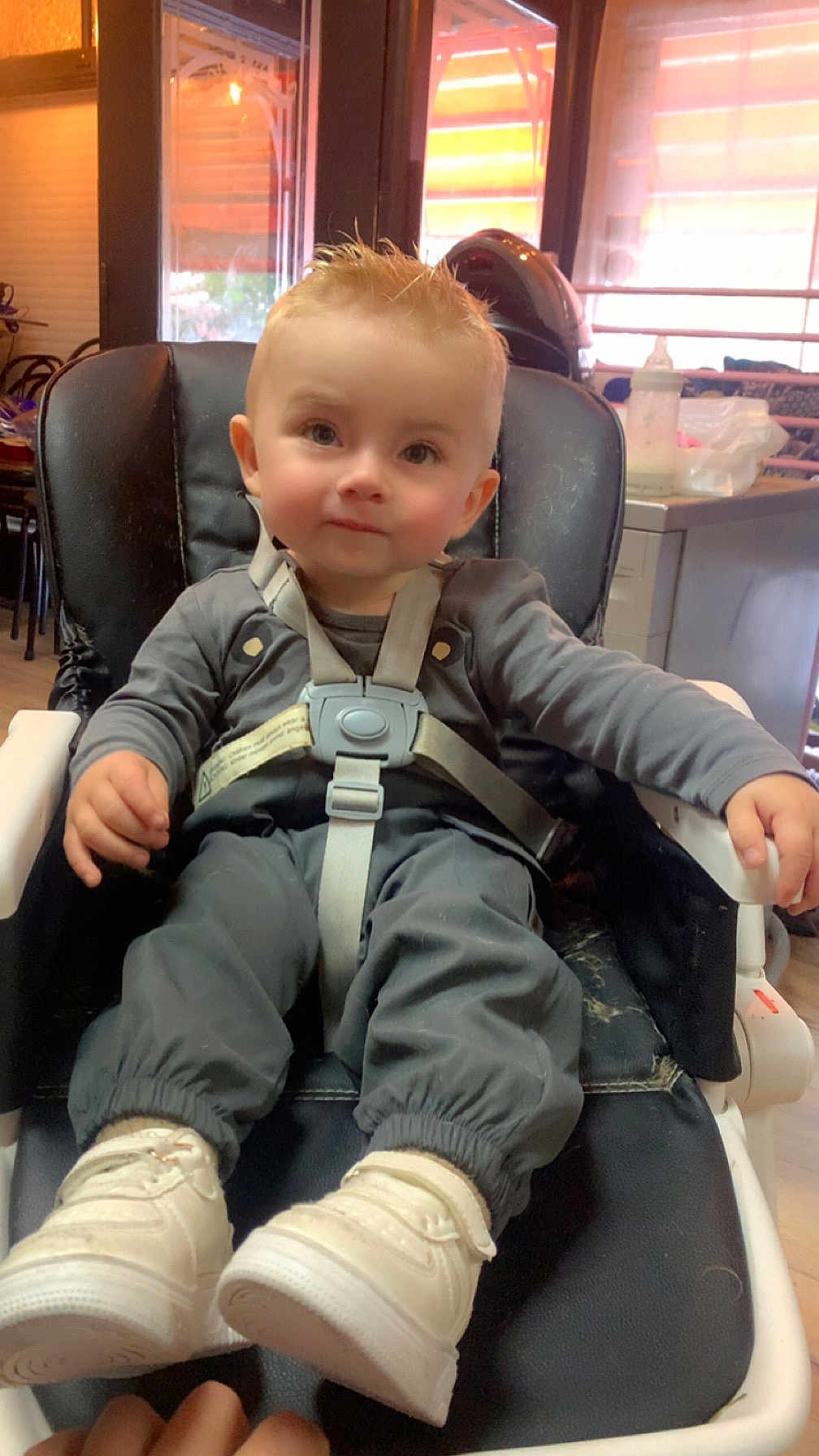Thymaël a rejoint le concours — aidez-le/la à gagner de superbes lots ! toddler, child, baby, high_chair, straps, white_shoes, gray_pants, long_sleeve_shirt, indoor, window, baby_bottle, furniture, seat, curious, face, hair, hands, floor, light, expression