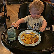 Thymaël participe au concours pour gagner de l'argent avec cette photo : toddler, child, high_chair, pasta, fork, sippy_cup, mickey_mouse, curly_hair, indoor, wooden_floor, feeding, plate, baby, eating, clothing, person, food, tray, chair, stroller