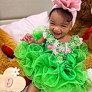 Amirah joined the competition — help win amazing prizes! baby, child, green_dress, headband, flower, smiling, teddy_bear, plush_toy, bed, pillow, milestone_sign, cute, happy, infant, sitting, portrait, celebration, indoors, fabric, accessory