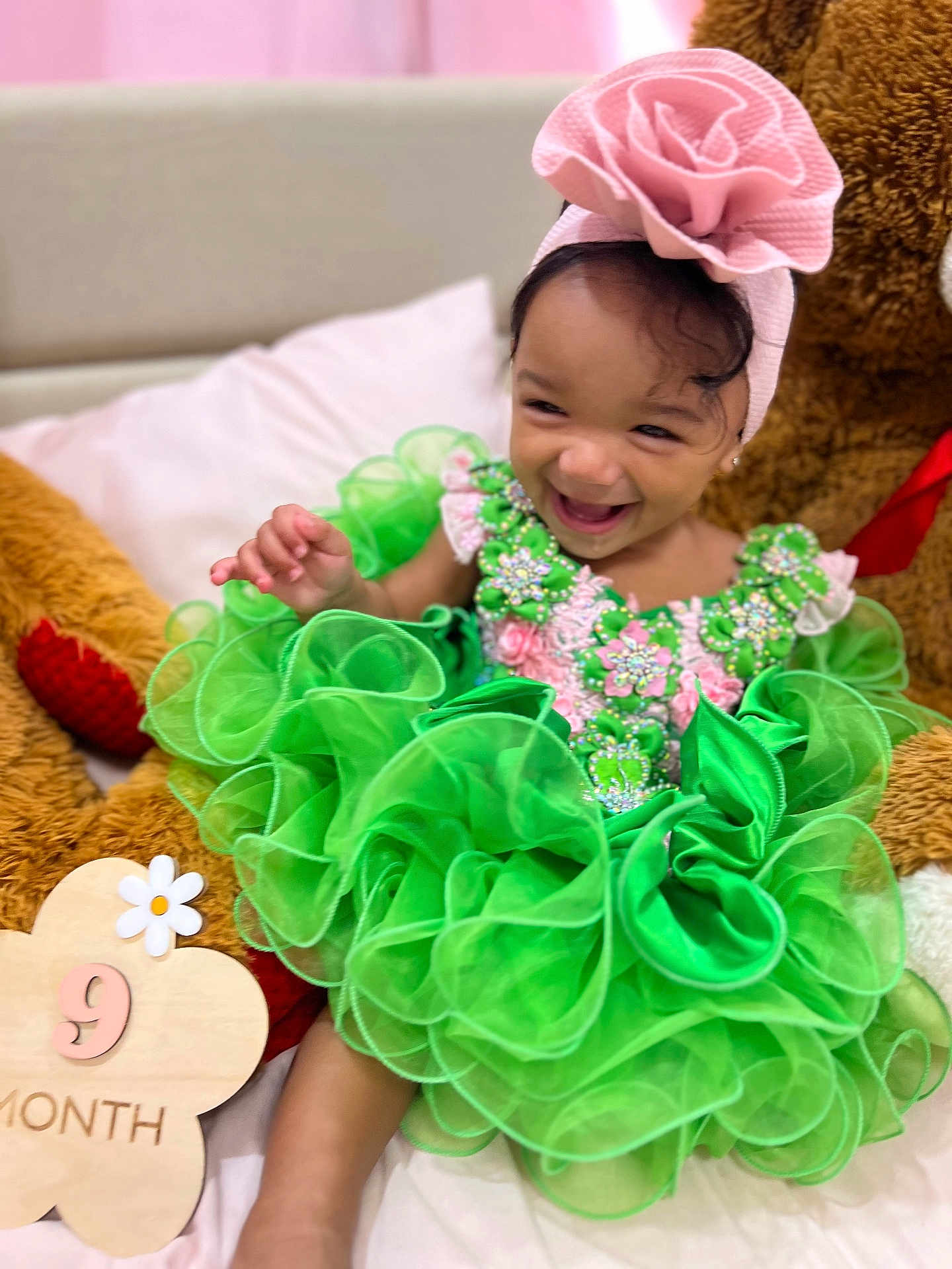 Amirah joined the competition — help win amazing prizes! baby, child, green_dress, headband, flower, smiling, teddy_bear, plush_toy, bed, pillow, milestone_sign, cute, happy, infant, sitting, portrait, celebration, indoors, fabric, accessory