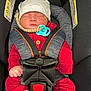 baby, sleeping, car_seat, red_clothing, white_hat, pacifier, beaded_clip, safety_harness, infant, child, cute, cozy, seatbelt, car_interior, warning_label, hands, headwear, resting, newborn, toddler