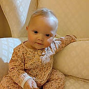 Asher is registered to the contest to win money with this photo: baby, child, infant, onesie, floral_pattern, couch, furniture, indoor, seated, cute, face, skin, head, hand, expression, comfort, soft_lighting, drool, curious, young