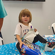 Kyler is registered to the contest to win money with this photo: child, toy, unwrapping, table, blue_tablecloth, white_tshirt, black_pants, shoes, indoor, television, wrapping_paper, gift, party, smiling, sitting, person, hair, face, floor, chair