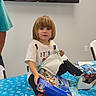 child, toy, unwrapping, table, blue_tablecloth, white_tshirt, black_pants, shoes, indoor, television, wrapping_paper, gift, party, smiling, sitting, person, hair, face, floor, chair