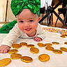 Rylan joined the competition — help win amazing prizes! baby, child, green_bow, headband, white_outfit, gold_coins, chocolate_coins, blanket, cereal, floor, indoors, smiling, happy, playful, hand, face, eyes, tongue, table, chairs