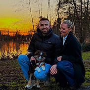 Nala participe au concours pour gagner de l'argent avec cette photo : couple, dog, puppy, outdoor, sunset, water, trees, smiling, grass, nature, jacket, jeans, footwear, happy, park, warm_clothing, evening, pets, people, crouching