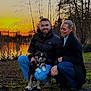 couple, dog, puppy, outdoor, sunset, water, trees, smiling, grass, nature, jacket, jeans, footwear, happy, park, warm_clothing, evening, pets, people, crouching