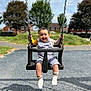 child, toddler, swing, playground, smiling, outdoor, daylight, grass, trees, houses, clothing, socks, happy, baby, seat, metal_chains, shadow, pavement, fun, nature