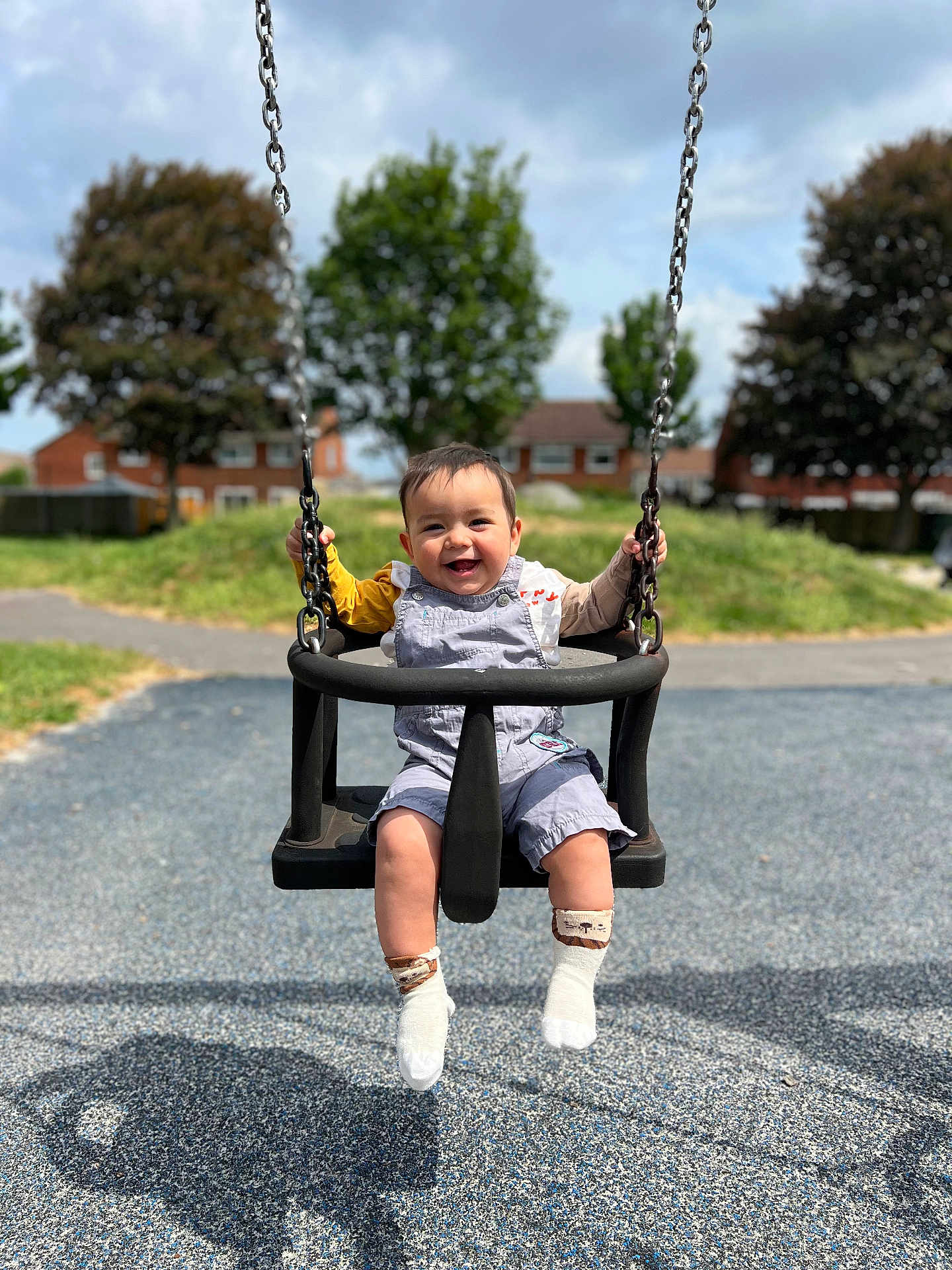 Mason is registered to the contest to win money with this photo: child, toddler, swing, playground, smiling, outdoor, daylight, grass, trees, houses, clothing, socks, happy, baby, seat, metal_chains, shadow, pavement, fun, nature