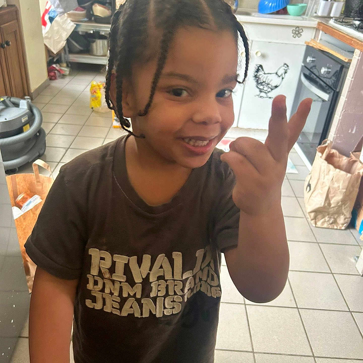 Soann participe au concours pour gagner de l'argent avec cette photo : child, smiling, braided_hair, hand_gesture, kitchen, tile_floor, cabinet, oven, paper_bag, vacuum_cleaner, dishware, person, indoor, appliance, countertop, food_packaging, holding_hands, casual_clothing, home_interior, playful