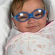 Jaxxon is registered to the contest to win money with this photo: baby, infant, glasses, blue_glasses, swaddle, blanket, pattern, heart_pattern, arrow_pattern, smile, face, head, cute, child, person, eyewear, soft_lighting, indoors, closeup, portrait