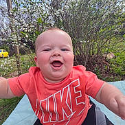 Easton is registered to the contest to win money with this photo: arms, baby, blanket, blossom, cheeks, face, flowering_bush, grass, happy, infant, nike_logo, outdoor, portrait, red_shirt, shorts, sitting, smiling, sunny, trees, yellow_car