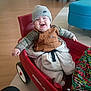 baby, infant, smiling, red_wagon, wagon, beanie, hat, overalls, blanket, indoor, wood_floor, child, toy, wheel, seat, happy, playful, cute, portrait, person