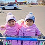 twin_babies, baby, pink_outfit, pom_pom_hat, shopping_cart, parking_lot, outdoor, white_car, stop_sign, mcdonalds, pavement, cloudy_sky, blanket, smiling, neutral_expression, winter_clothing, infant_seat, retail_store, portrait, cute