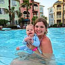 woman, baby, swimming_pool, water, smile, happy, outdoor, sunny, resort, vacation, family, child, holding, building, palm_tree, summer, people, blue_sky, playful, relaxation