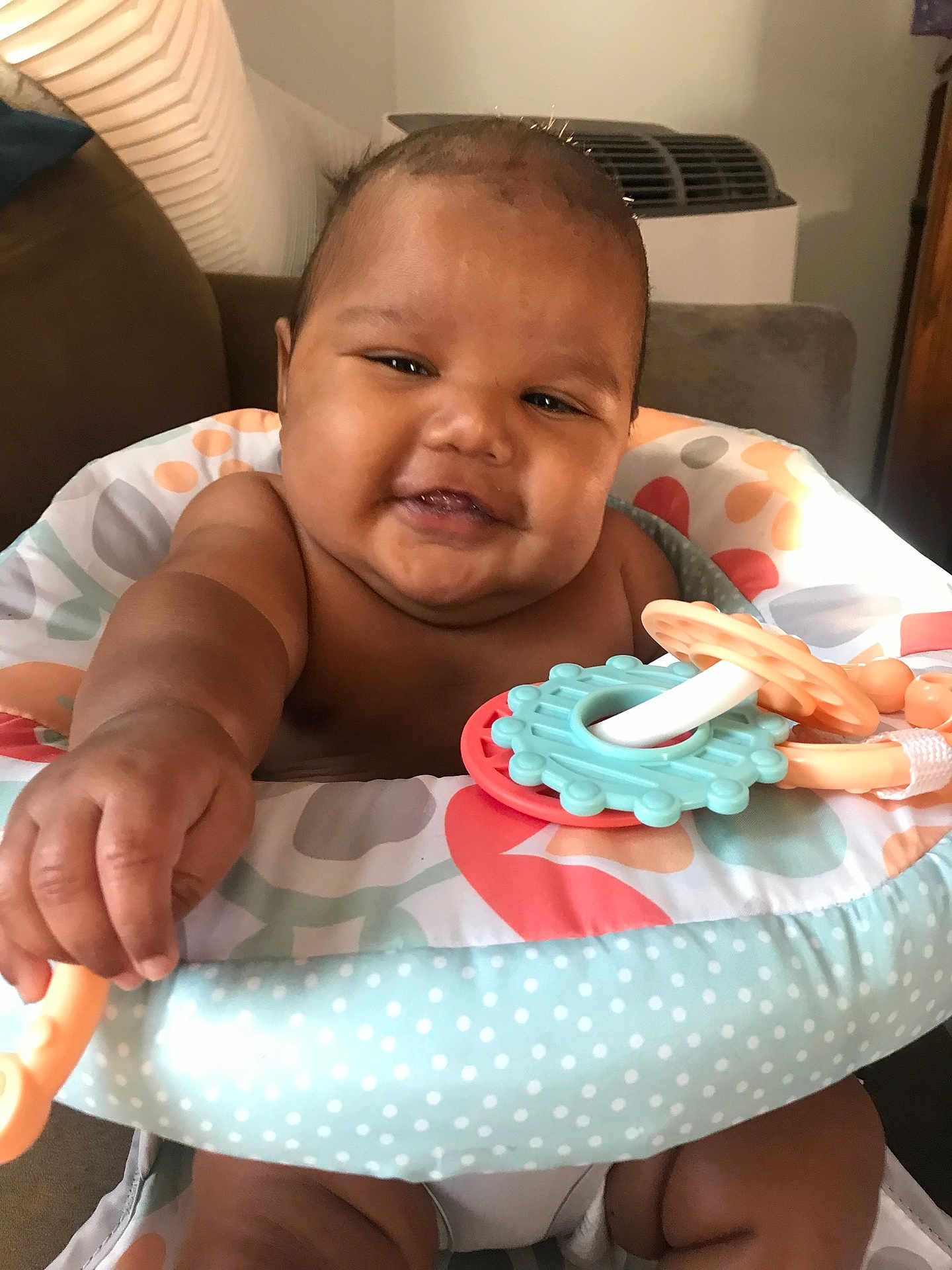 Daniel is registered to the contest to win money with this photo: baby, child, infant, smiling, happy, teething_toy, colorful, polka_dot, baby_seat, arm, hand, face, cheeks, indoors, furniture, couch, living_room, skin, cute, playful