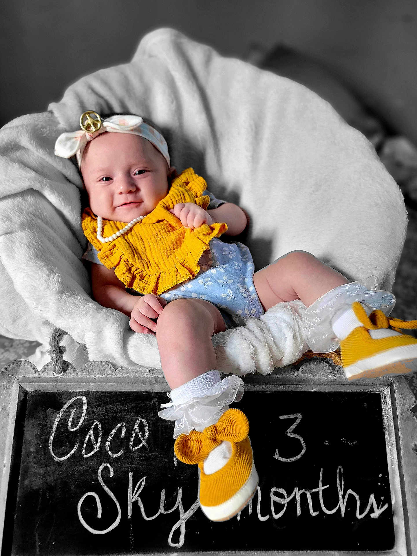 Coco is registered to the contest to win money with this photo: baby, infant, smiling, headband, yellow_bib, blue_onesie, white_chair, fluffy_cushion, pearl_necklace, yellow_shoes, chalkboard, milestone, portrait, cute, cozy, indoor, child, happy, sitting, decor