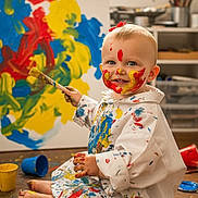 Tommie-Rose joined the competition — help win amazing prizes! toddler, child, paint, paintbrush, canvas, art, messy, face_paint, floor, studio, creative, colorful, white_smock, smiling, blue_paint, yellow_paint, red_paint, barefoot, indoor, playful