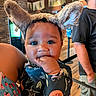 baby, child, infant, portrait, face, smiling, happy, bunny_ears, costume, hand, fingers, dark_clothing, patterned_clothing, restaurant, indoor, adult, background, wood_floor, seated, close_up