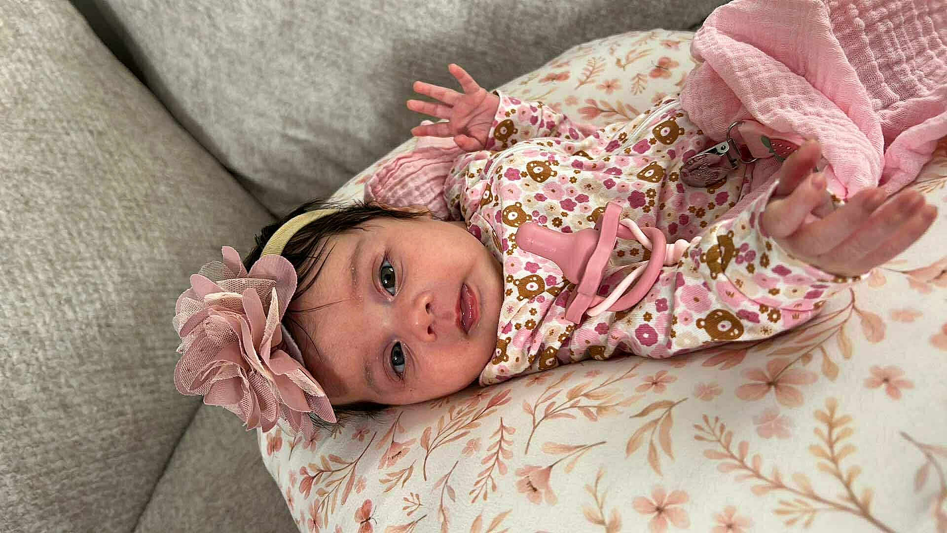 Sadie is registered to the contest to win money with this photo: baby, infant, flower_headband, pacifier, pink, cushion, floral_pattern, clothing, hand, face, eyes, cute, lying_down, comfort, indoors, child, newborn, soft, head, person