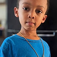 Messiah is registered to the contest to win money with this photo: child, boy, portrait, indoors, standing, blue_shirt, cartoon_shirt, necklace, short_hair, curly_hair, serious_expression, looking_at_camera, face, upper_body, hands, clothing, home, background, photography, close_up