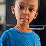 child, boy, portrait, indoors, standing, blue_shirt, cartoon_shirt, necklace, short_hair, curly_hair, serious_expression, looking_at_camera, face, upper_body, hands, clothing, home, background, photography, close_up