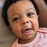 baby, blanket, child, close_up, cozy, curly_hair, cute, dark_skin, eyes, face, floral_pattern, happy, indoor, infant, lying_down, onesie, pink_clothing, portrait, smiling, tongue_out