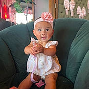 Emerson is registered to the contest to win money with this photo: baby, child, infant, headband, flower, pink, pacifier, green_chair, indoor, decor, wall_bows, cute, smiling, sitting, clothing, dress, toes, happy, person, portrait