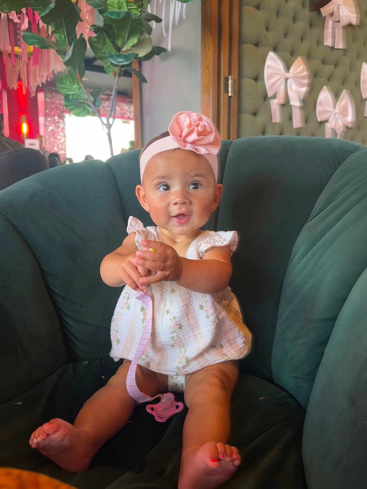 Emerson is registered to the contest to win money with this photo: baby, child, infant, headband, flower, pink, pacifier, green_chair, indoor, decor, wall_bows, cute, smiling, sitting, clothing, dress, toes, happy, person, portrait