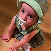 Kolter is registered to the contest to win money with this photo: baby, child, pacifier, hat, green_clothing, sitting, wooden_floor, indoor, cute, face, hands, looking_up, floor, infant, one_person, toddler, casual, expression, young_child, portrait