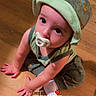 baby, child, pacifier, hat, green_clothing, sitting, wooden_floor, indoor, cute, face, hands, looking_up, floor, infant, one_person, toddler, casual, expression, young_child, portrait