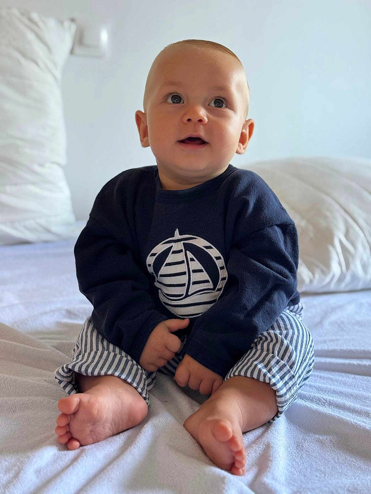 Louis participe au concours pour gagner de l'argent avec cette photo : baby, child, infant, sitting, bed, blue_clothing, striped_pants, barefoot, indoors, soft_light, cute, portrait, curious, face, hands, feet, pillow, blanket, home, casual