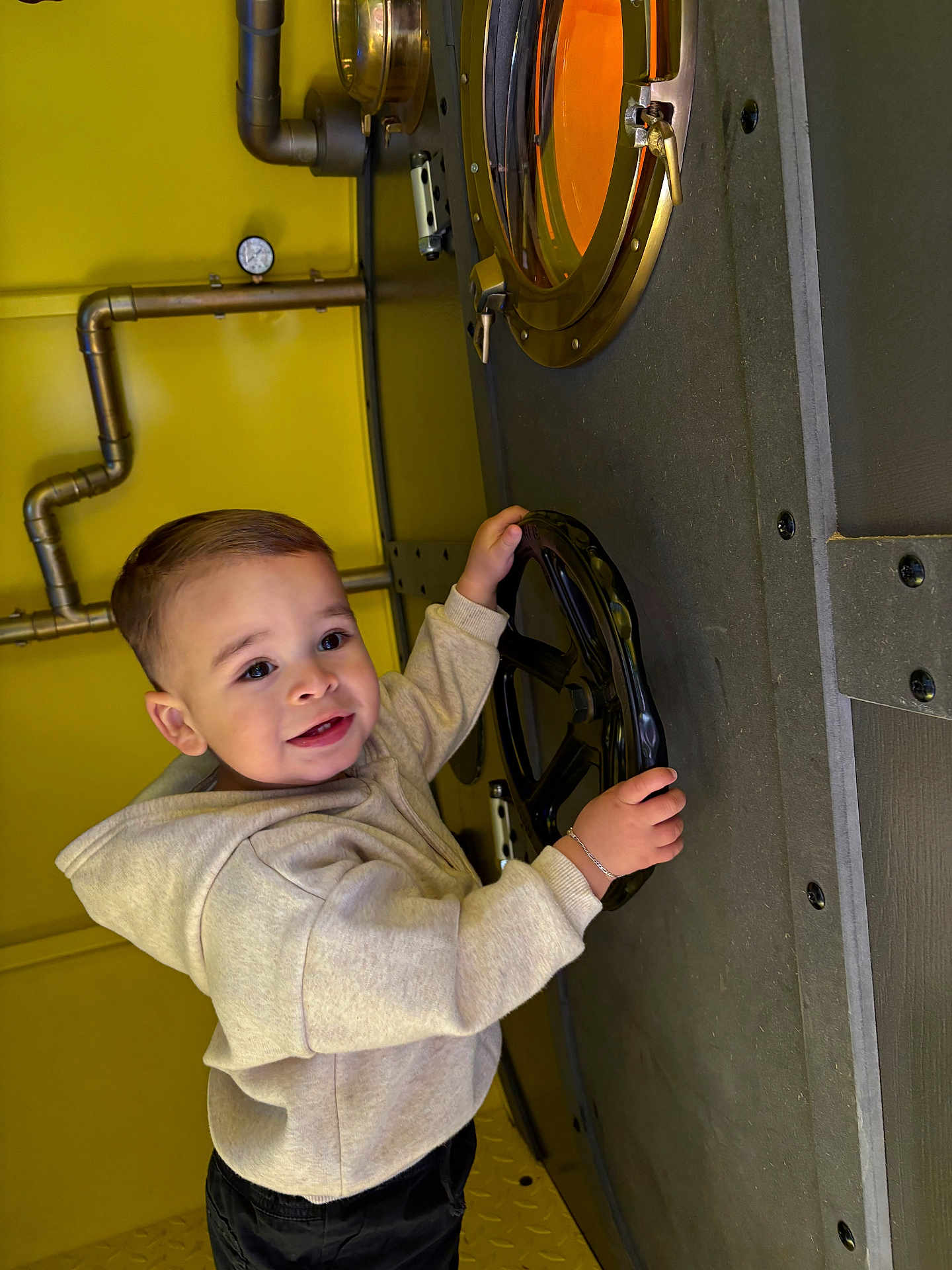 Kaïs a rejoint le concours — aidez-le/la à gagner de superbes lots ! toddler, child, hoodie, black_pants, wheel, industrial, yellow_wall, metal, pipe, gauge, curious, smiling, short_hair, bracelet, indoor, playful, young_child, person, casual_clothing, exploring