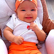 Mikanto participe au concours pour gagner de l'argent avec cette photo : baby, infant, smile, headband, bow, orange_pants, white_shirt, baby_seat, cushion, happy, cute, child, indoors, portrait, person, clothing, seatbelt, skin, hands, face