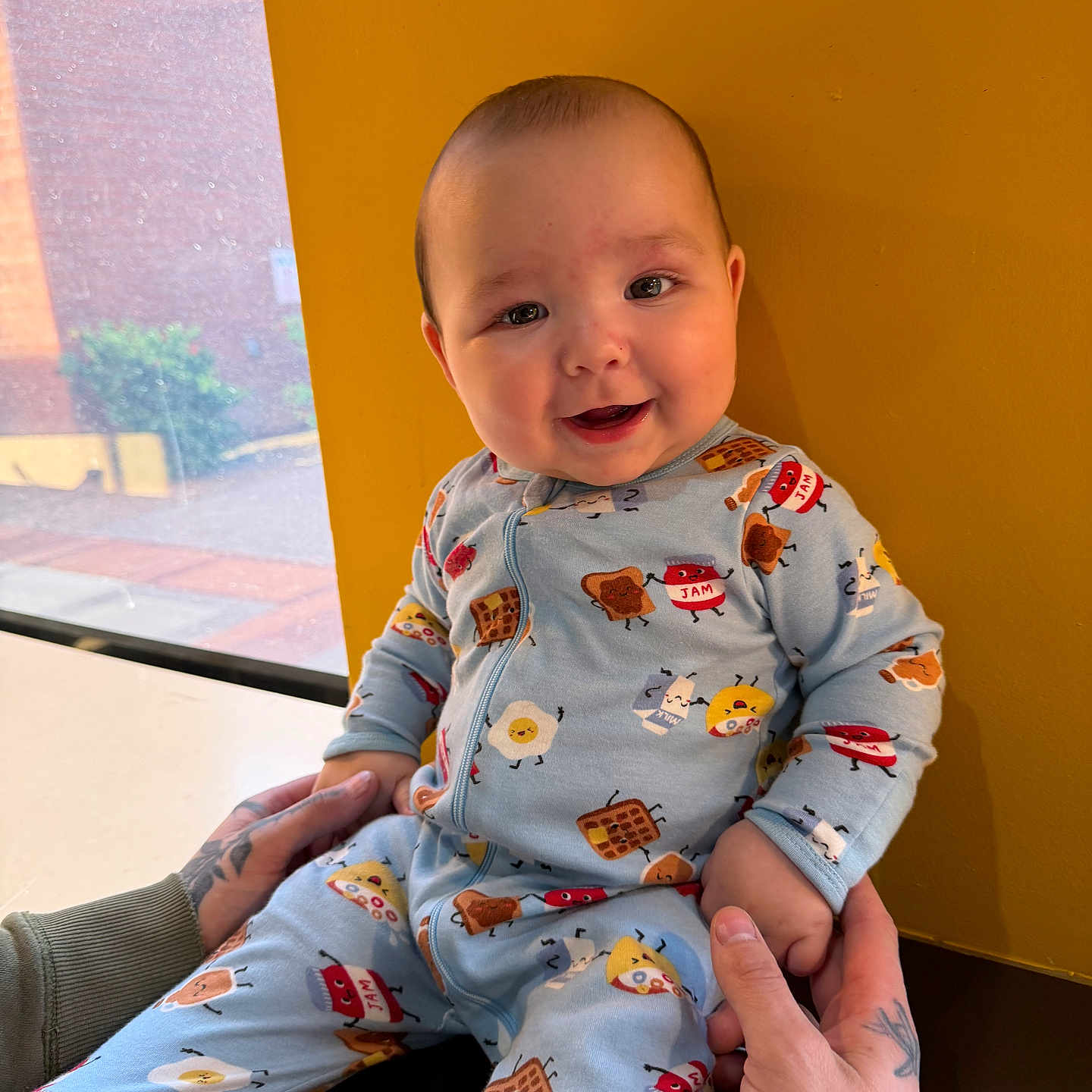 Andre is registered to the contest to win money with this photo: baby, child, smiling, onesie, tattoo, hands, window, yellow_wall, indoor, seated, cute, portrait, infant, playful, happy, face, clothing, person, background, casual