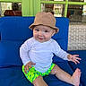 baby, child, smiling, hat, bucket_hat, white_shirt, green_shorts, floral_pattern, blue_cushion, sitting, indoor, window, reflection, sunlight, playful, happy, person, foot, hand, casual_clothing