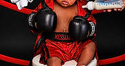 Messiah is registered to the contest to win money with this photo: baby, boxing_gloves, red_trunks, stool, milk_bottle, towel, child, portrait, studio, black_background, boxing_robe, serious_expression, feet, hands, cross_necklace, sportswear, infant, cute, preparation, indoor