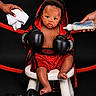 baby, boxing_gloves, red_trunks, stool, milk_bottle, towel, child, portrait, studio, black_background, boxing_robe, serious_expression, feet, hands, cross_necklace, sportswear, infant, cute, preparation, indoor