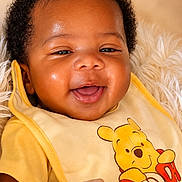 Messiah is registered to the contest to win money with this photo: baby, smiling, infant, yellow_bib, winnie_the_pooh, cute, portrait, child, happy, face, closeup, soft_texture, warm_colors, curly_hair, clothing, indoors, adorable, young_child, cute_expression, joyful
