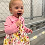 casual_clothing, chain_link_fence, child, cute, daylight, floral_dress, food, girl, happy, lollipop, metal_bleachers, outdoor, pink_cardigan, playful, portrait, smiling, standing, sweet, toddler, young_child